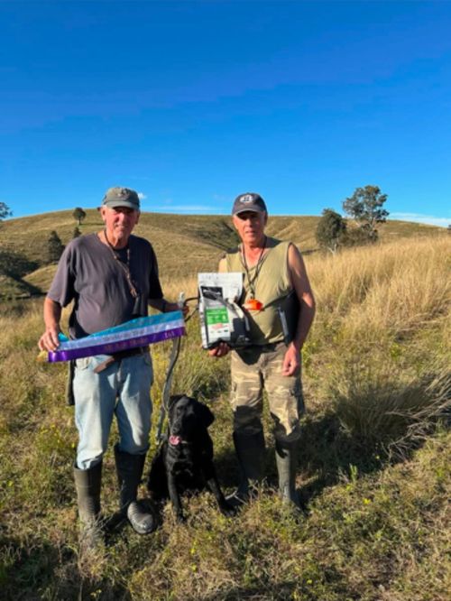 Flash, a black Labrador Retriever, with two handlers holding awards in a grassy field